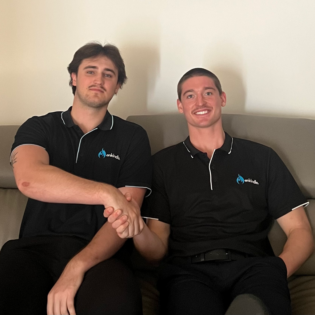 Two individuals, identified as Harvie and Kobi, the co-founders and co-directors of Enkindle Marketing & Management, are seen seated together in black polo shirts with the company logo, exuding professionalism and camaraderie in a relaxed setting